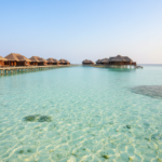 Calm turquoise lagoon and wooden overwater villa boardwalk in the Maldives under a bright blue sky.