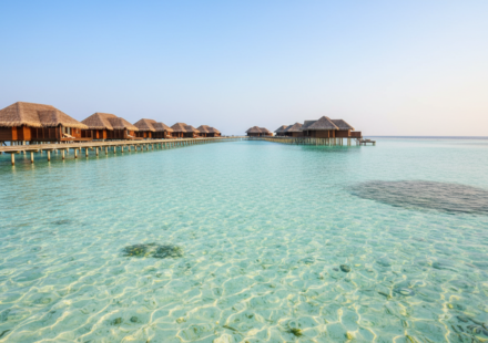 Calm turquoise lagoon and wooden overwater villa boardwalk in the Maldives under a bright blue sky.