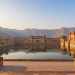 Sandstone walls of Amber Fort overlooking a calm lake surrounded by misty hills at sunrise in Jaipur.