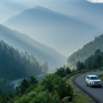 A white SUV drives along a winding mountain road through misty green valleys in North East India.