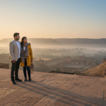 A couple stands on a sandstone terrace at sunrise overlooking historic forts and distant mist-covered hills.