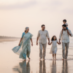 An Indian multi-generational family strolling along a quiet Goan beach at sunset with calm ocean waves.