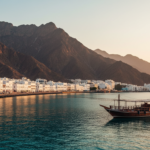 White Omani coastal buildings and mountains along Mutrah Corniche in Muscat under a clear golden hour sky.