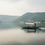 A wooden boat on calm Lake Pichola in Udaipur surrounded by green Aravalli hills under soft overcast skies.