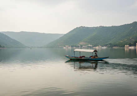 A wooden boat on calm Lake Pichola in Udaipur surrounded by green Aravalli hills under soft overcast skies.