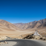 A winding road through the arid, sunlit mountains of Ladakh under a clear blue August sky.