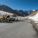 A paved Himalayan mountain road in Ladakh surrounded by melting snow walls and sharp barren rock peaks under a clear blue sky.