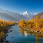 Golden autumn trees along a clear blue river with snow-capped mountain peaks in the background under a blue sky.