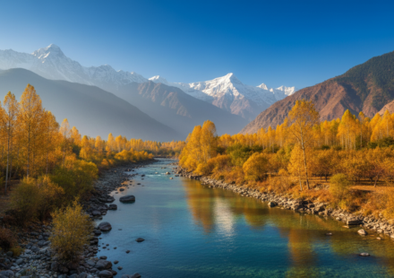 Golden autumn trees along a clear blue river with snow-capped mountain peaks in the background under a blue sky.