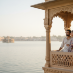Couple on a heritage hotel balcony overlooking Lake Pichola in Udaipur at morning golden hour.