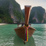 A traditional wooden longtail boat anchored in clear turquoise water near dramatic limestone cliffs in Thailand.