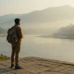A traveler with a backpack stands by the Ganges River in Rishikesh surrounded by green hills during morning mist.