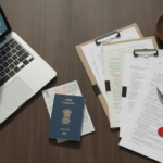 Overhead flat-lay of an Indian passport, travel documents, bank statements, and a laptop on a wooden desk.
